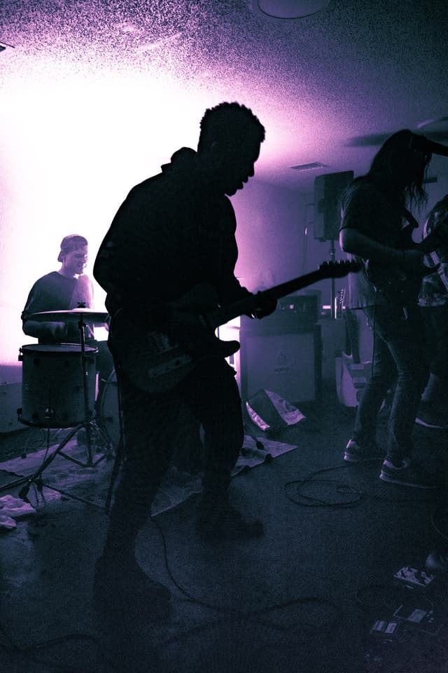 A Live Alone guitarist silhouetted against pale violet stage light, with the drummer behind him
